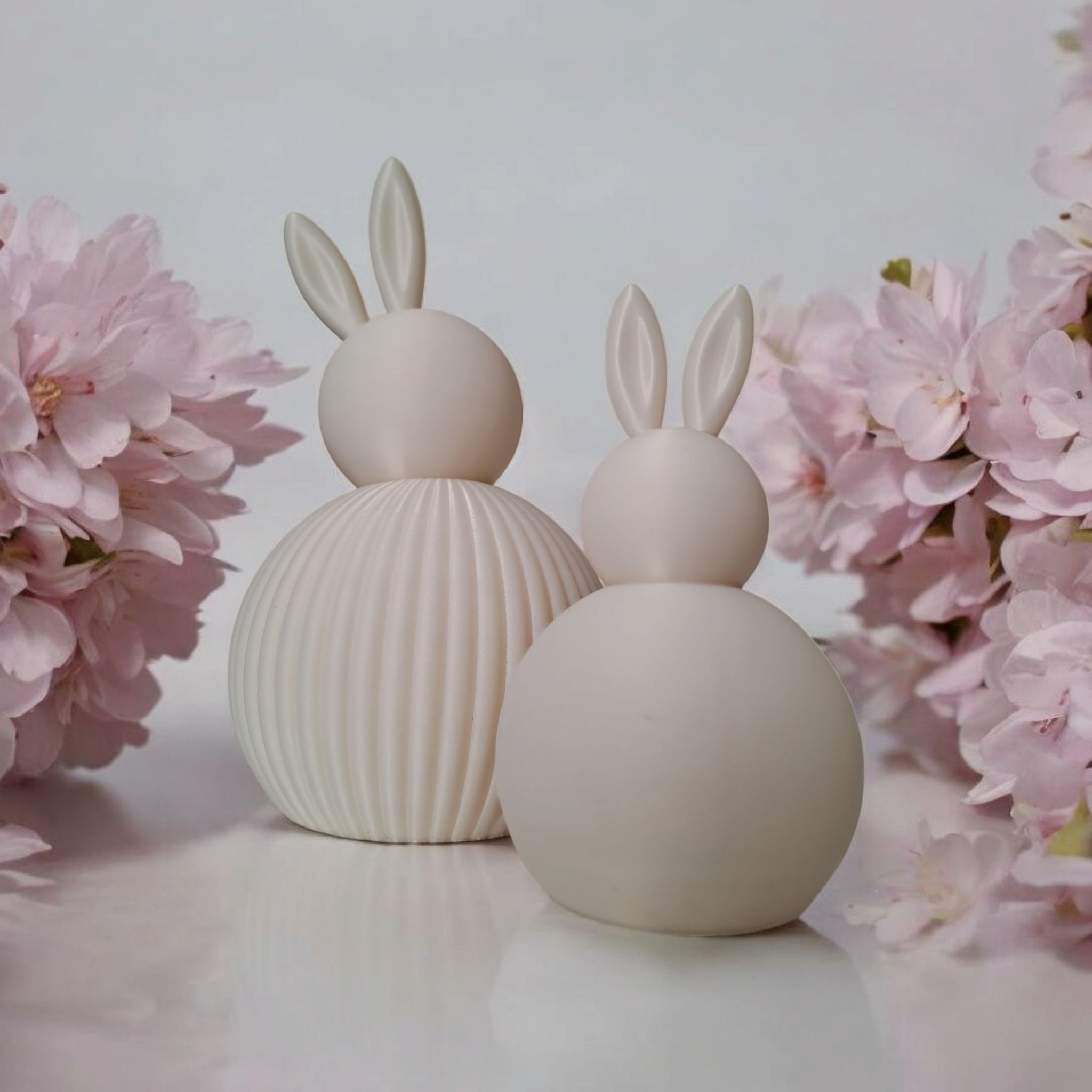 To hvide 3D printede påskeharer (rillet og glat) opstillet sammen ved siden af kirsebærblomster.