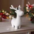 Hvid 3D printet rensdyr figur stående på lys træbord med julepynt og lyskæde i baggrunden.