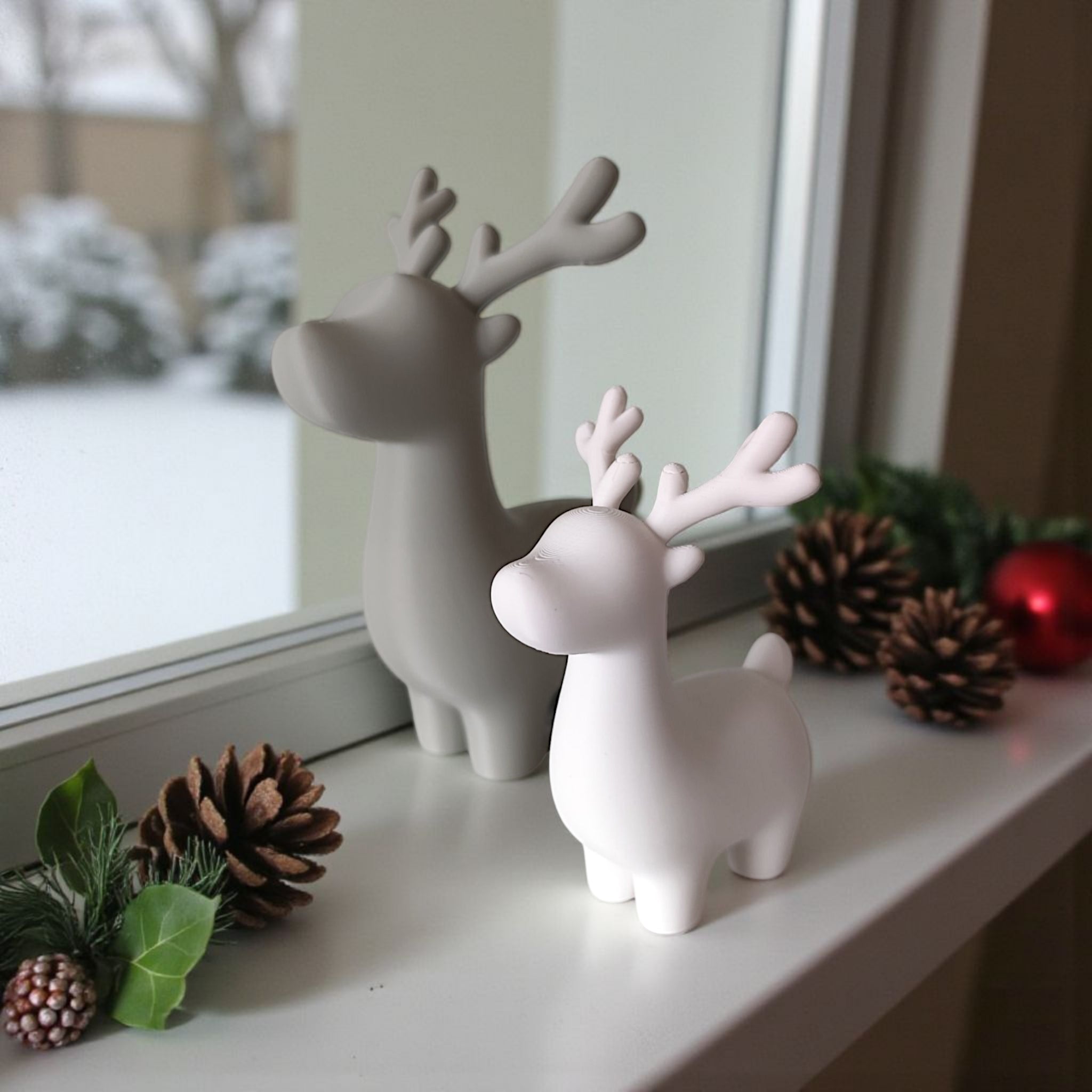 To 3D printede rensdyr figurer i grå og hvid på vindueskarm med kogler og gran, mens sne ses udenfor.