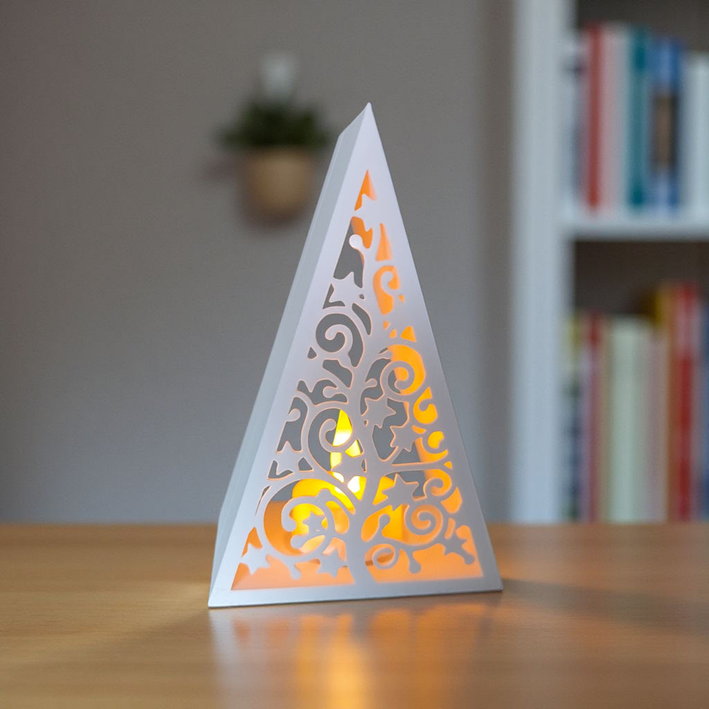 Minimalistisk hvid 3D-printet juletræs-lanterne med et gennembrudt, hvirvlende mønster, oplyst indefra af et varmt, gult LED-lys. Står på et træbord.