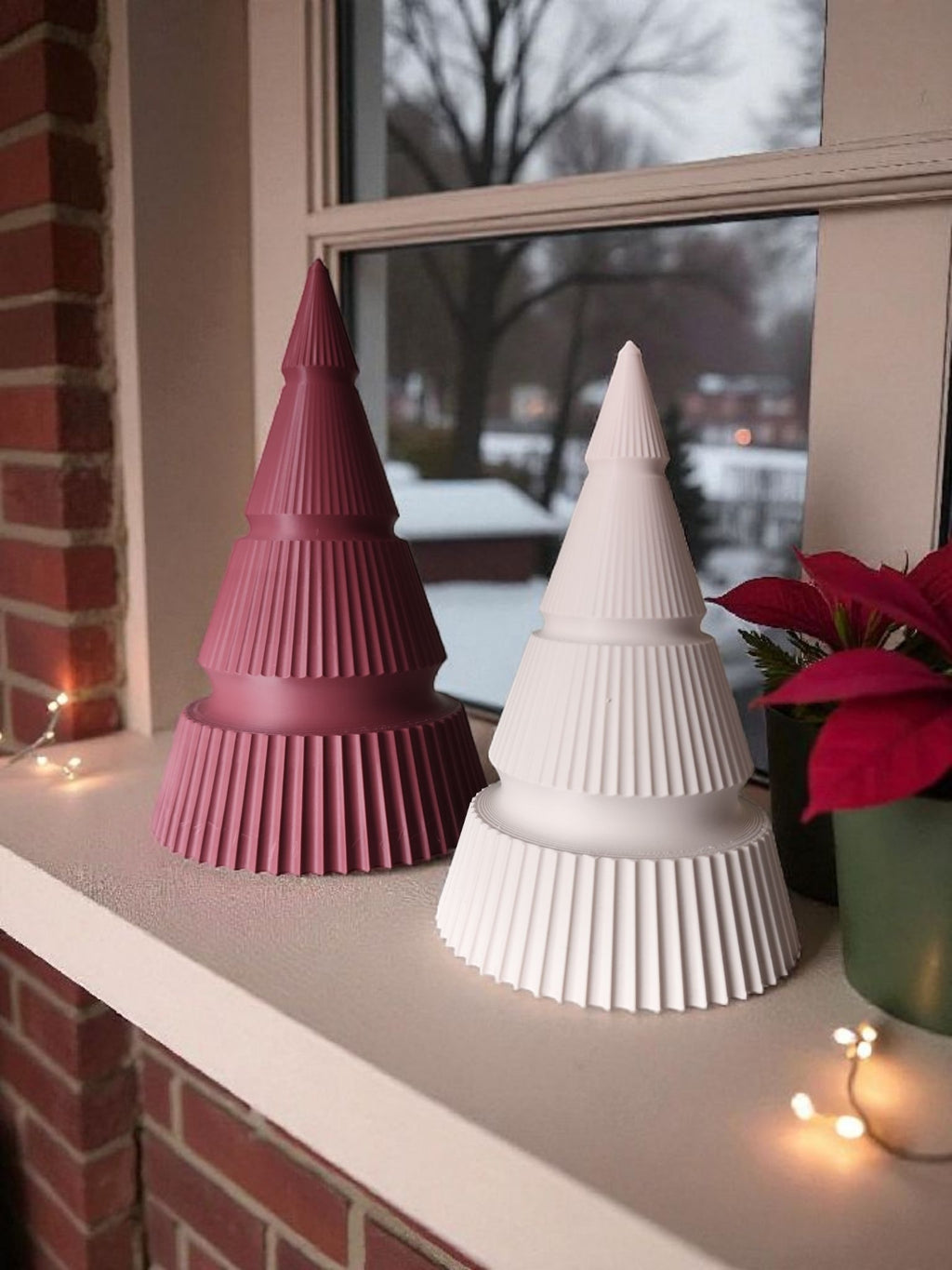 To 3D printede moderne juletræer i røde og hvide farver, placeret i en vindueskarm ved siden af LED lys, en plante og udsigt til sne i baggrunden.