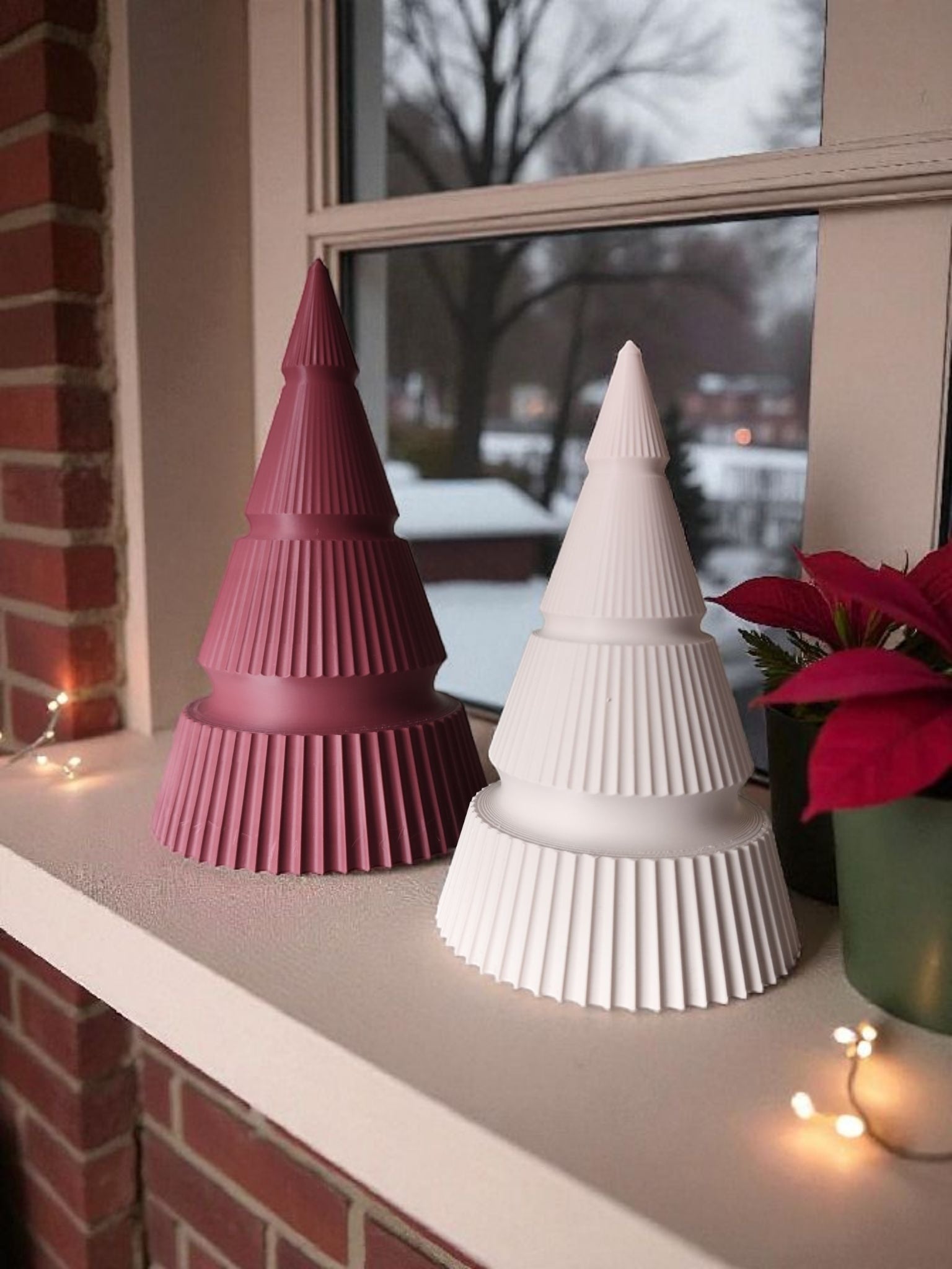 To 3D printede moderne juletræer i røde og hvide farver, placeret i en vindueskarm ved siden af LED lys, en plante og udsigt til sne i baggrunden.